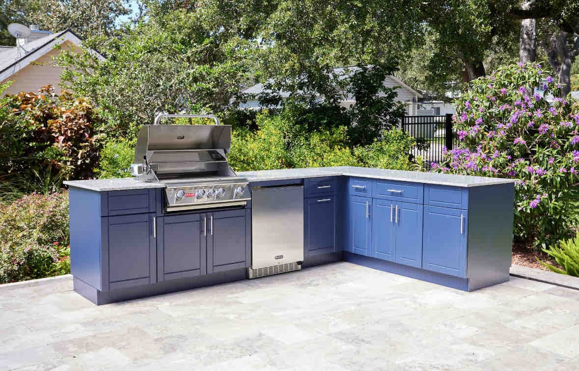 A Symphony Blue and Galaxy White DuraBULL outdoor kitchen on a stone patio in a backyard full of trees, bushes and flowers.