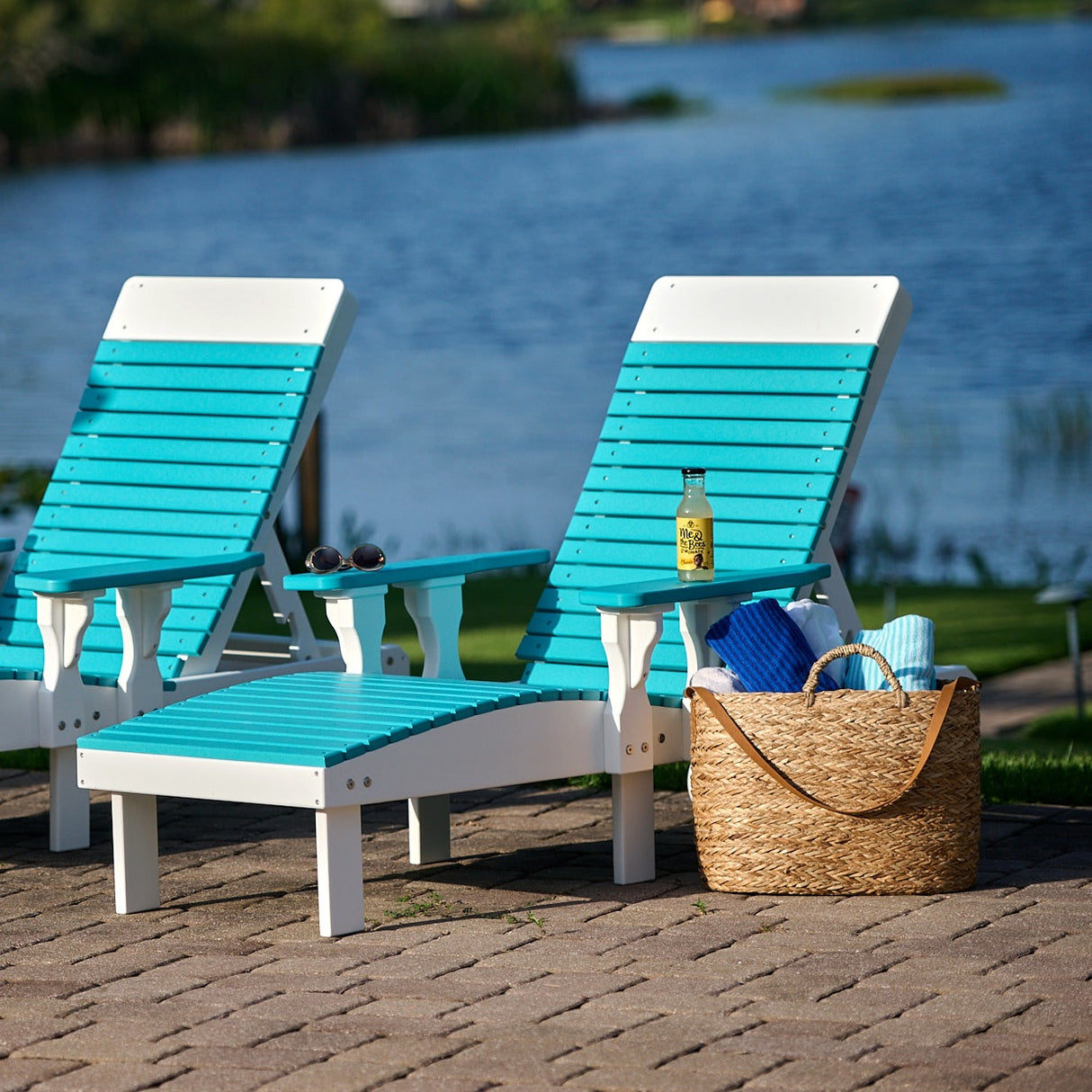 This is a lifestyle image showing two Aruba Blue and White colored lounge chairs on a brick patio in front of a body of water.