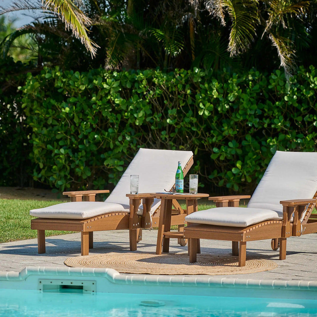 This is a lifestyle image showing two Antique Mahogany colored lounge chairs on a patio in front of a pool. There are lounge chair cushions on the chairs (those are sold separately).