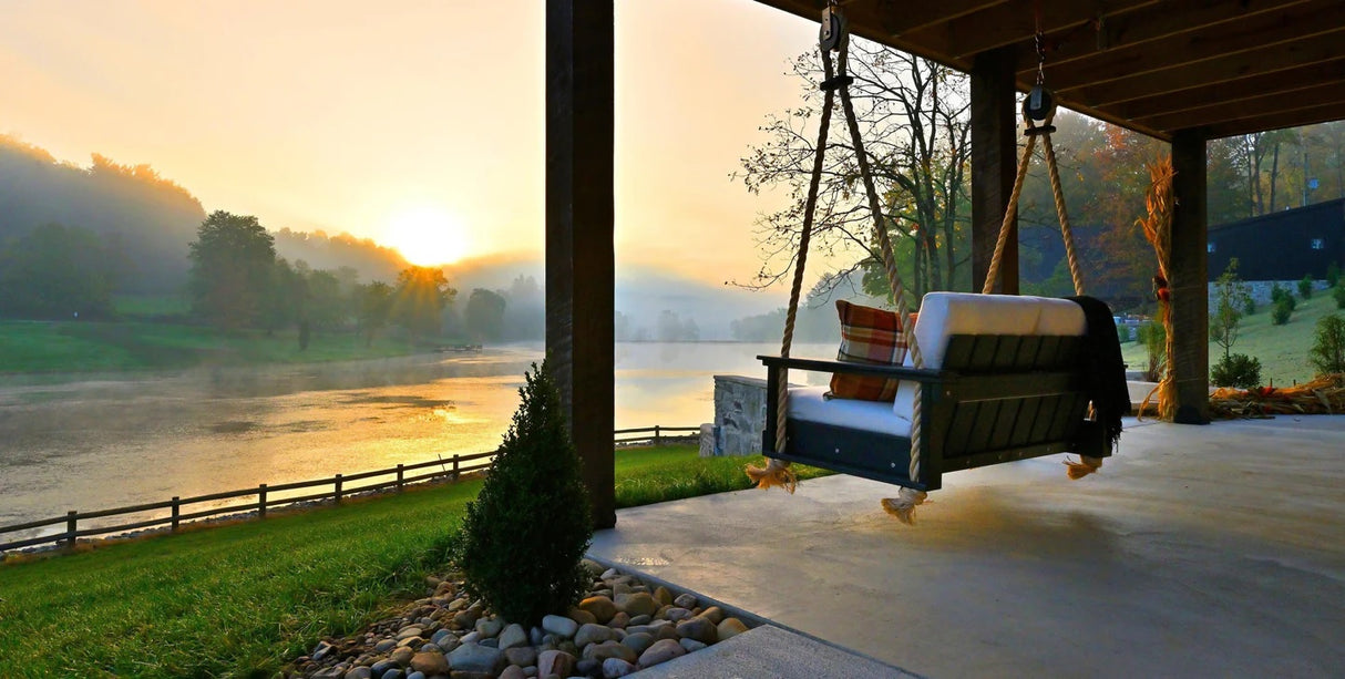 This is a lifestyle image showing the Lanai Loveseat Swing from behind. It's hanging from the roof of an outdoor patio facing a beautiful sunset over a large body of water.
