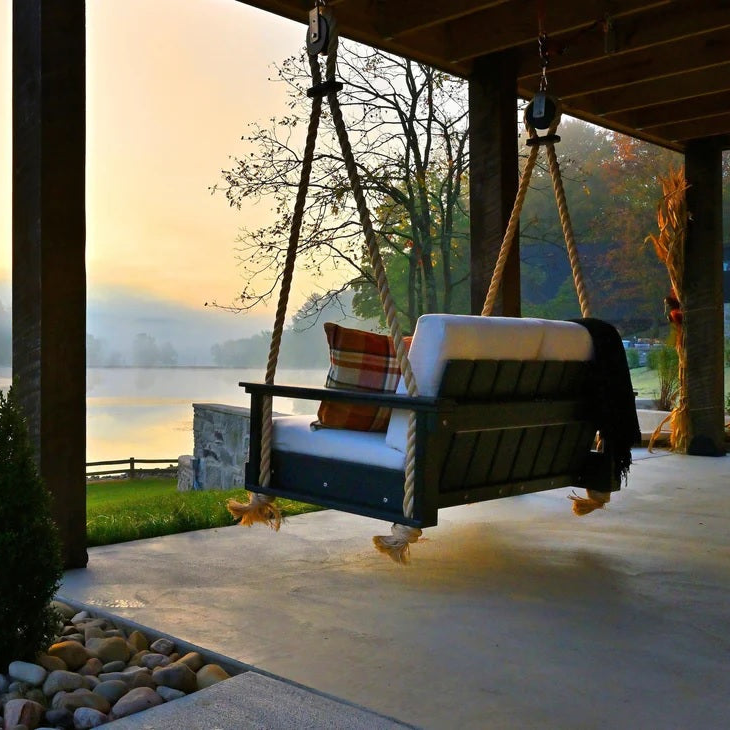 This is a lifestyle image showing the Lanai Loveseat Swing from behind. It's hanging from the roof of an outdoor patio facing a beautiful sunset over a large body of water.