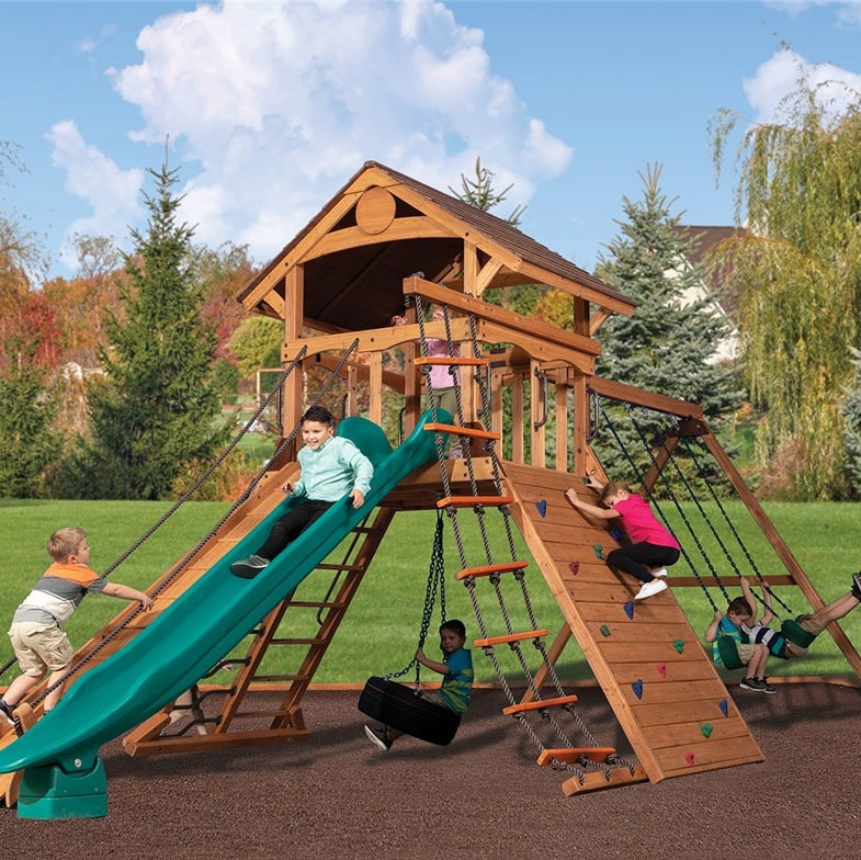 This is a lifestyle image showing this specific wooden playset in a grassy backyard. There's a boy using the rope ladder to climb up. There's a boy sliding down the green slide. There's a boy underneath the playset sitting on a tire swing. There's a little girl climbing the rock wall and two boys on the swings.