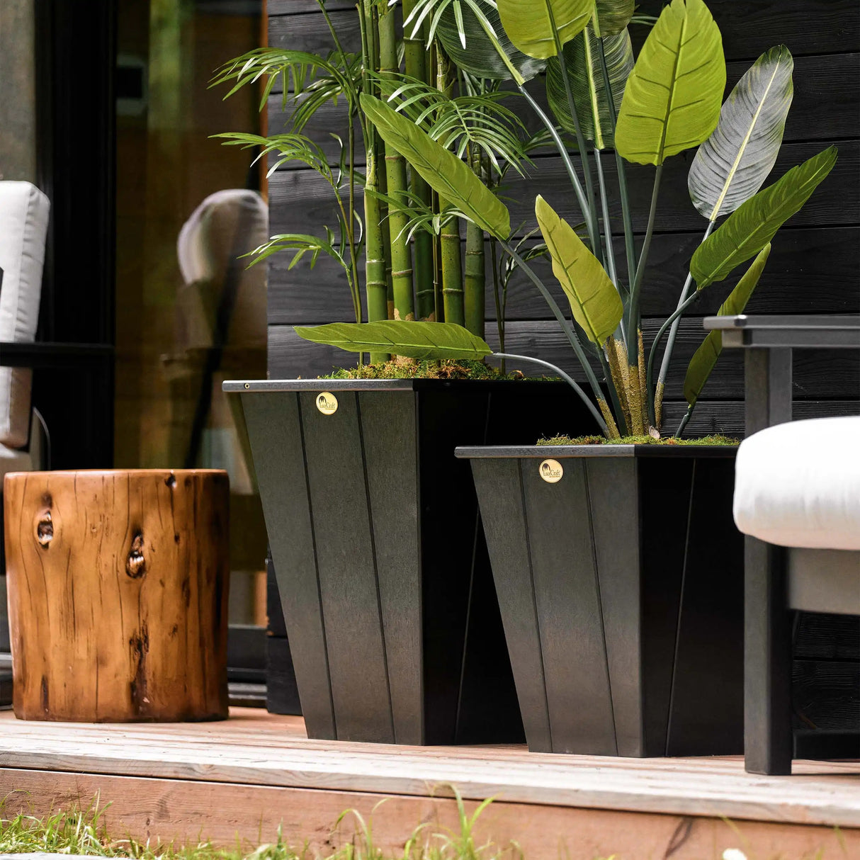 This is a lifestyle image showing two different sized Black planters on a wooden deck with plants growing inside them.