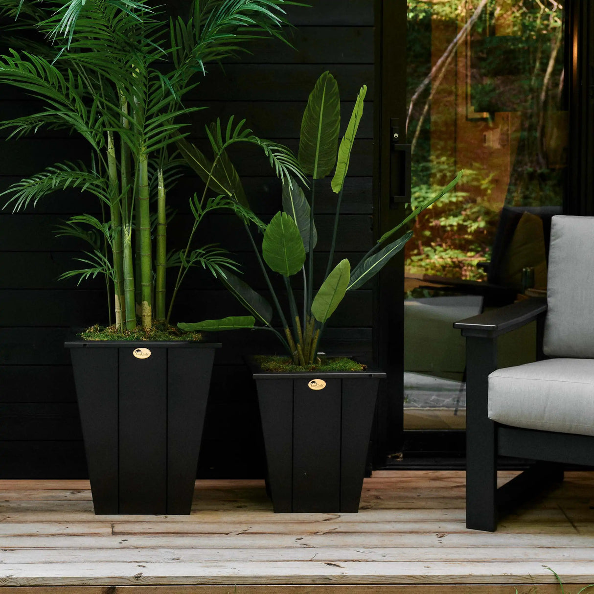 This is a lifestyle image showing two different sized Black colored planters on a wooden porch next to a cushioned patio chair.