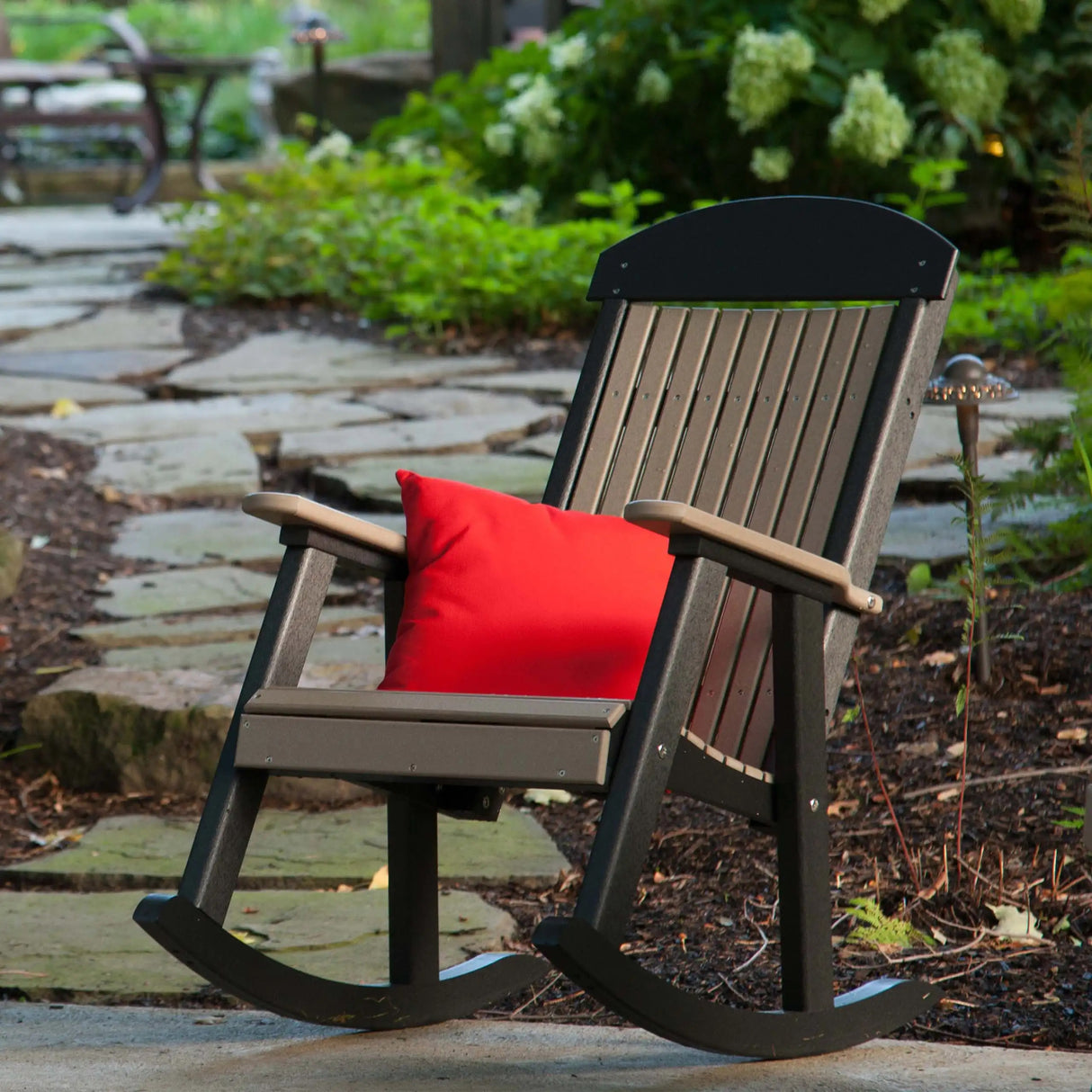 This is a lifestyle image showing a Weatherwood and Black colored porch rocker on a stone patio in a grassy backyard.
