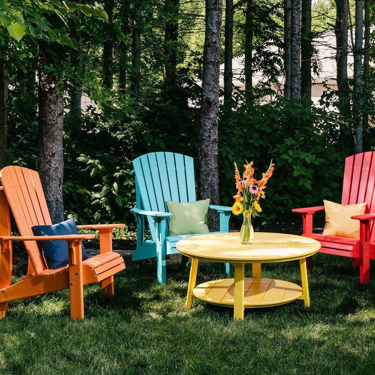 This is a lifestyle image showing three Adirondack chairs in the woods around a yellow table. The chair on the left is Tangerine, the chair in the middle of Aruba Blue and the chair on the right is Red.
