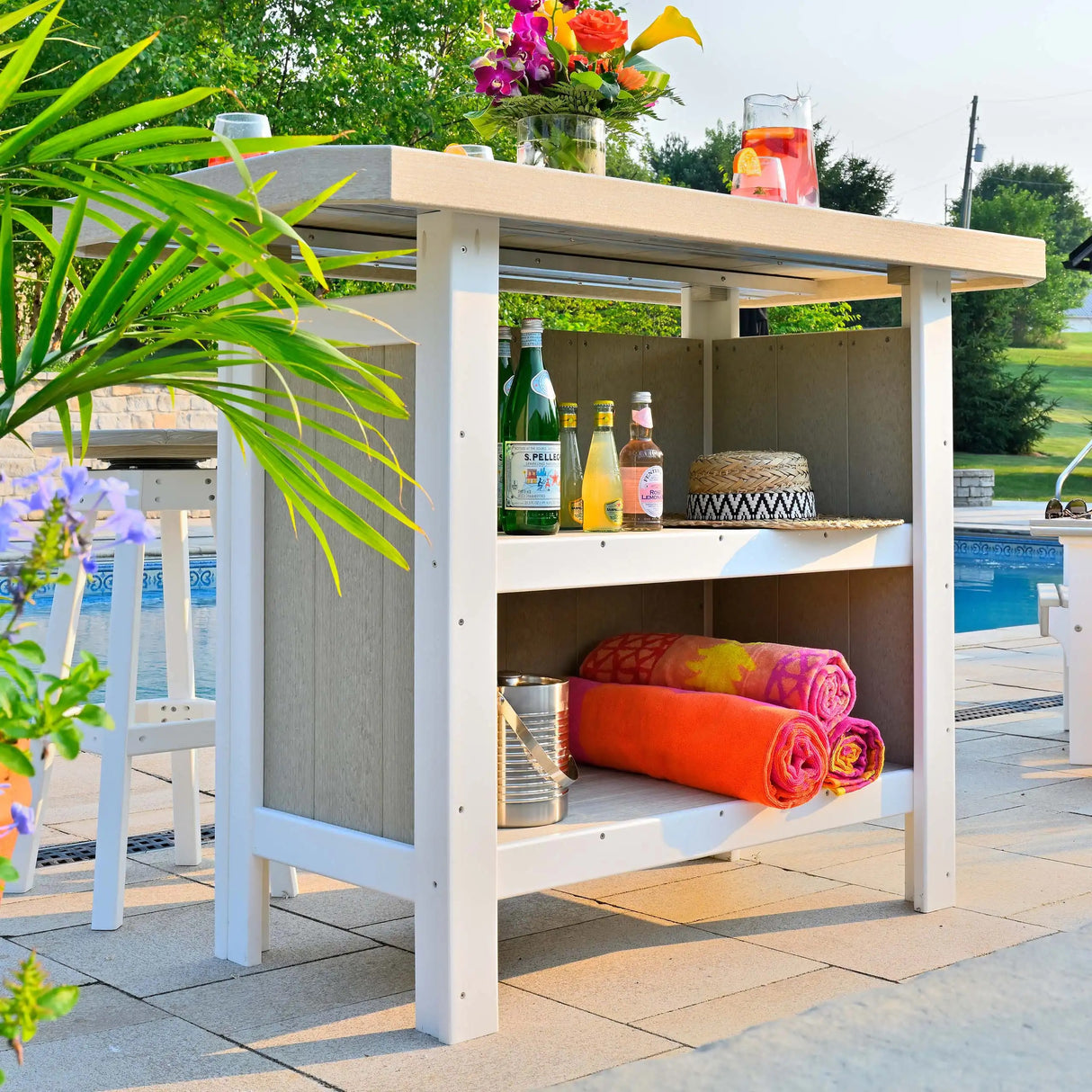 This is a lifestyle image showing the shelves inside the Birch and White colored Serving Bar. On the top of the Serving Bar is a pitcher of strawberry lemonade and a vase of pretty flowers. On the inside, on the top shelf, there are bottles of sparkling water and a straw hat. On the bottom shelf, there's a tin can and three beach towels.