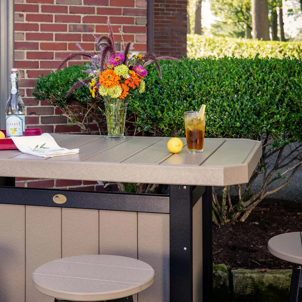This is a lifestyle image showing the Weatherwood and Black colored Serving Bar with two matching stools in front it. On top of the bar, there's a vase filled with pretty flowers, a glass of iced tea with a lemon wedge on the side and a glass of sparkling water.
