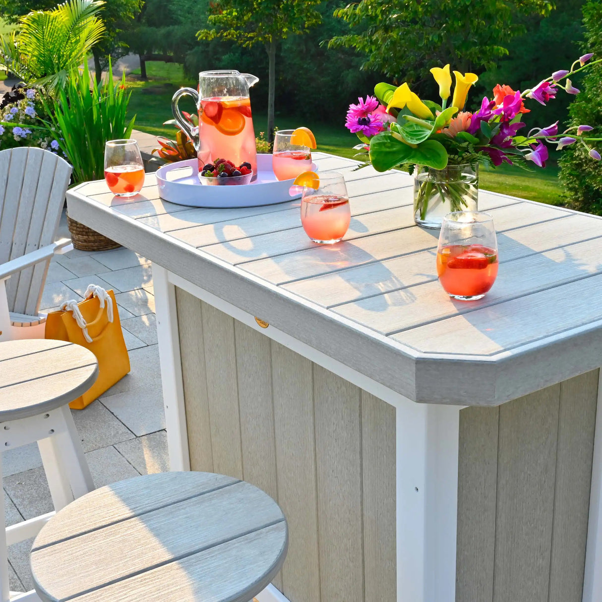 This is a lifestyle image showing the front of the Birch and White colored Serving Bar on an outdoor patio. There are two matching stools placed in front of the bar. On top of the bar is a pitcher of strawberry lemonade and four glasses full of fruit.