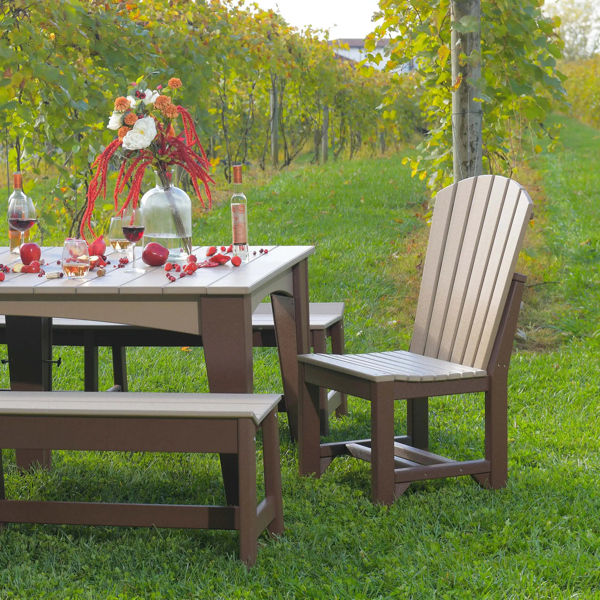 This is a lifestyle image showing the Weatherwood and Chestnut Brown colored Adirondack Side Chair in front of a matching table and benches. There are wine glasses, fruit and flowers on the table.