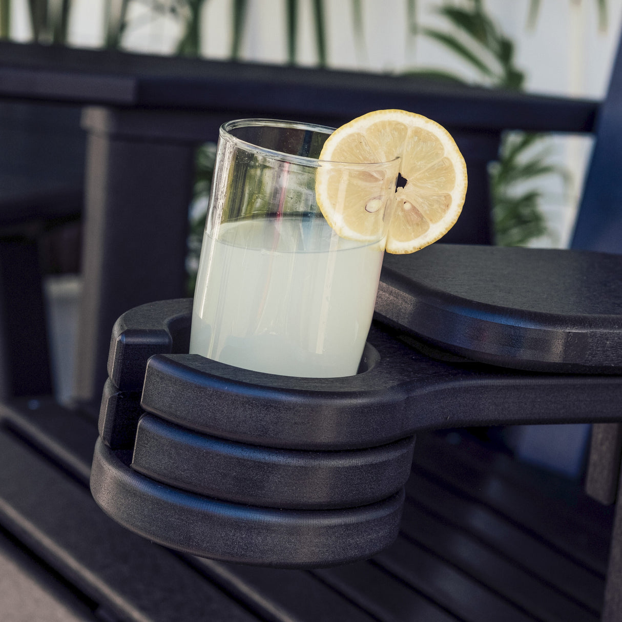 This is a lifestyle image showing a glass of lemonade inside the black colored cup holder that's attached to a black colored Adirondack chair.