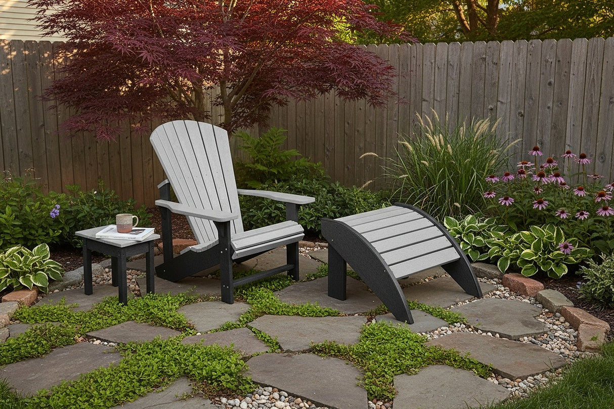 This is a lifestyle image showing the gray and black Adirondack chair and matching end table and ottoman on a stone and grass patio. There's a wooden fence and lots of pretty Midwestern foliage.