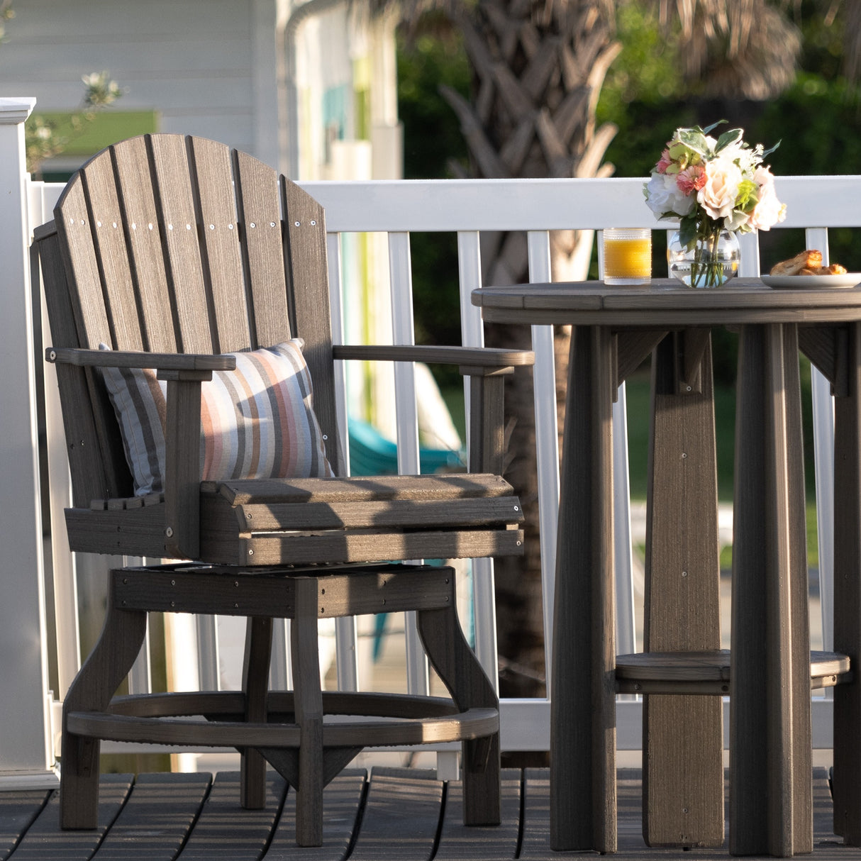 This is a lifestyle image showing the Coastal Gray colored swivel Adirondack chair next to a matching table on a wooden deck.