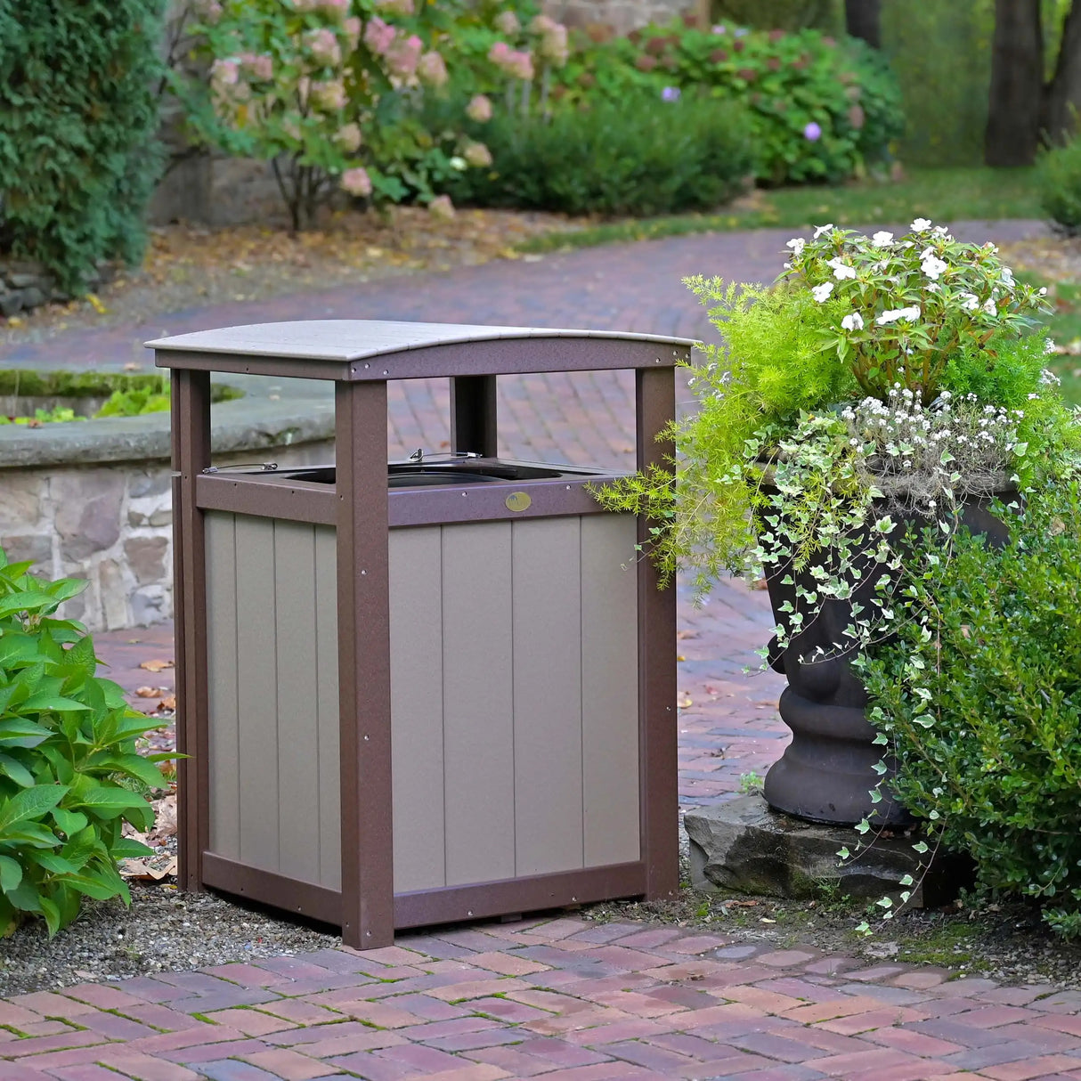 This is a lifestyle image showing the Weatherwood and Chestnut Brown colored garbage can on a brick patio surrounded by plants.