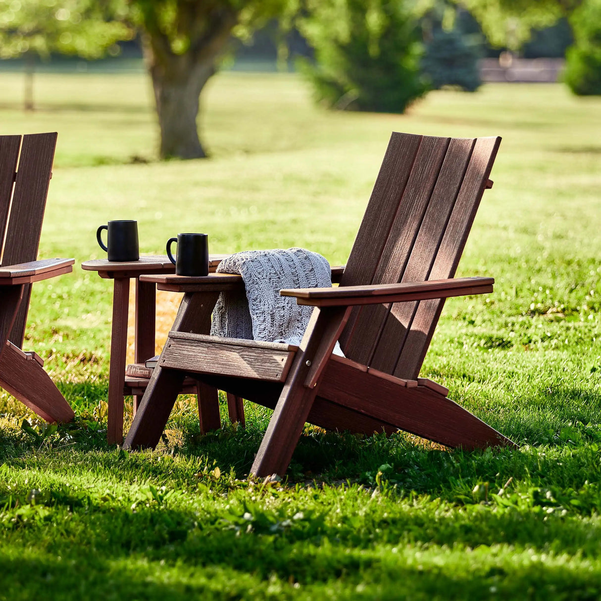 This is a lifestyle image showing the Antique Mahogany colored Adirondack chair next to a matching table in a grassy field.