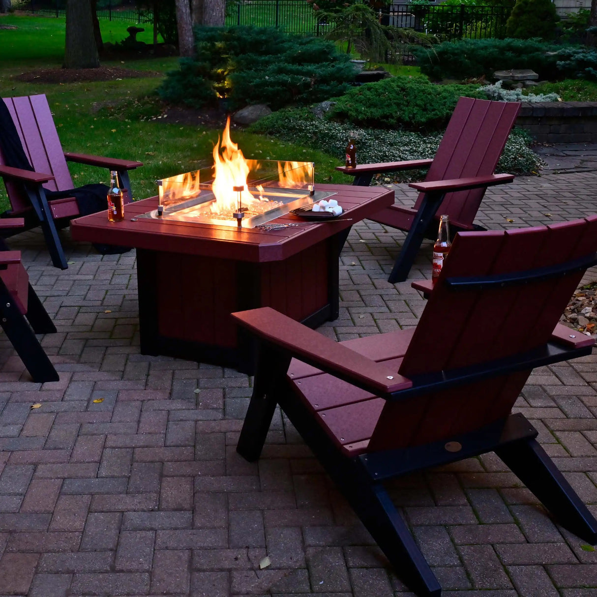This is a lifestyle image showing four Cherrywood and Black colored Adirondack chairs around a matching fire pit table on a brick patio.