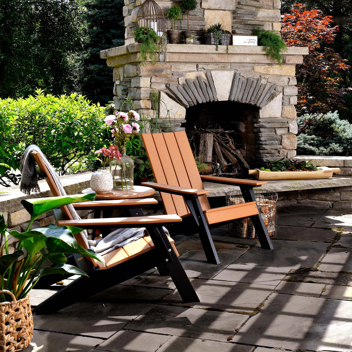 This is a lifestyle image showing two Cedar and Black colored Adirondack chairs on a stone patio next to an outdoor fireplace.