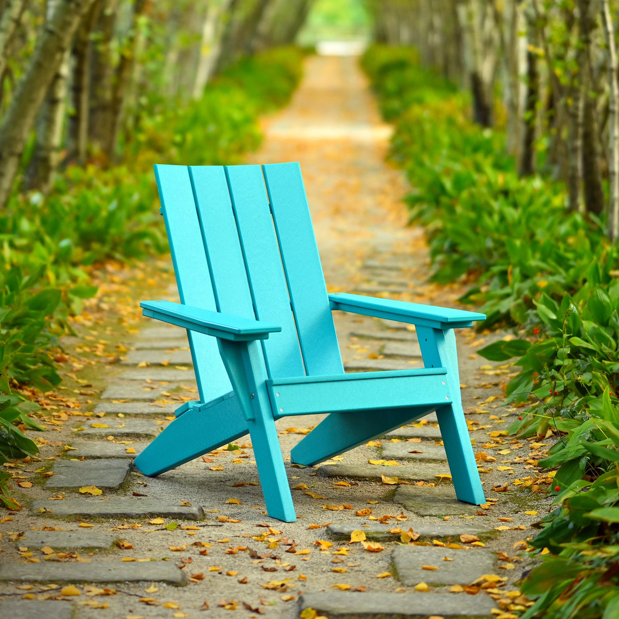 This is a lifestyle image showing the Aruba Blue colored Adirondack chair on a stone walkway surrounded by trees and plants.