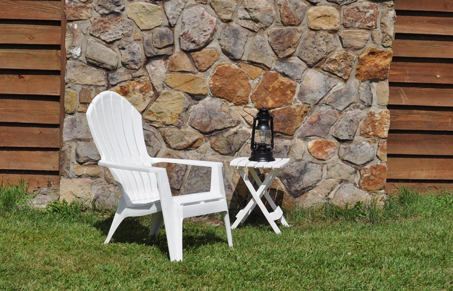 A white Adirondack chair outside on grass next to a stone chimney.