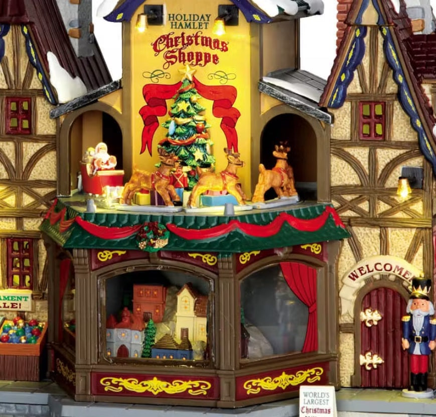 Porcelain lighted building for a Lemax display. Designed to look like a rustic cabin covered in snow and decorated for Christmas. There are angels and pigeons flying above. Santa is waving from a window in the attic. There are reindeer going in a circle around a decorated Christmas tree. There are ornaments for sale on the sidewalk and a nutcracker standing guard at the front door.