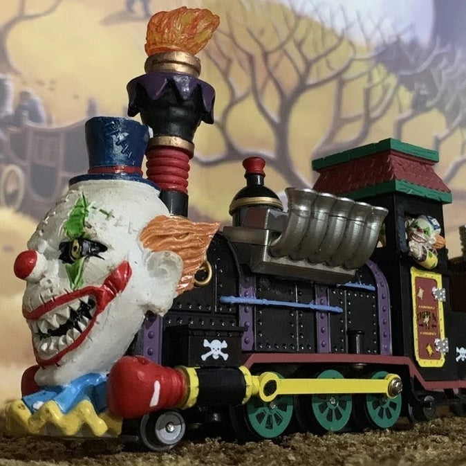 Polyresin train on a set of tracks for a Lemax display. The train cars are all different. The head of the train is the face of a clown. The second one is full of silver and wooden barrels and the side reads "Stitches the Clown: Traveling Mayhem." The third car is shaped like a giant clown shoe. The last one is shaped like a carnival ride where it spins you in a circle.