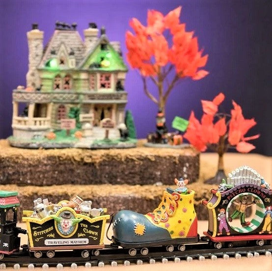 Polyresin train on a set of tracks for a Lemax display. The train cars are all different. The head of the train is the face of a clown. The second one is full of silver and wooden barrels and the side reads "Stitches the Clown: Traveling Mayhem." The third car is shaped like a giant clown shoe. The last one is shaped like a carnival ride where it spins you in a circle.
