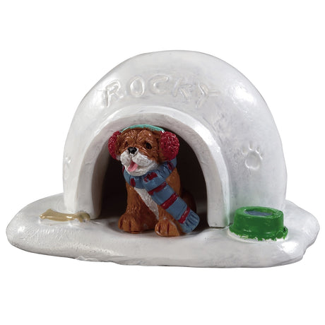 Polyresin figurine for a Lemax Village tabletop display. Designed to look like an igloo with a dog inside wearing earmuffs and a scarf. The name Rocky is written in the snow and there's a paw print on the side