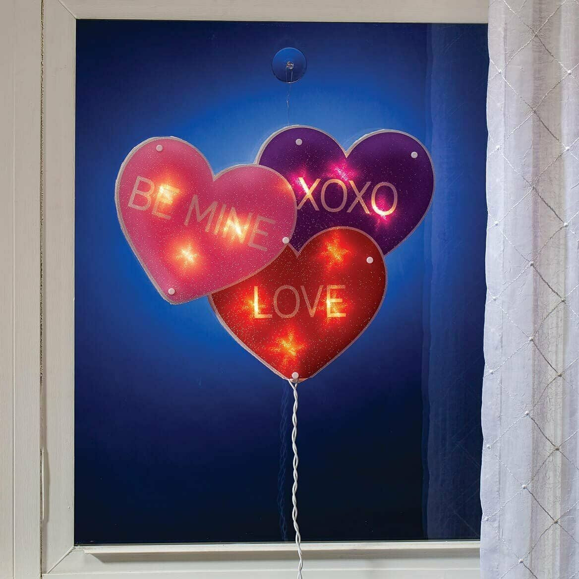 A light up decoration for Valentine's Day designed to look like three conversation hearts. One is purple and reads "XOXO." One is pink and reads "BE MINE." The other is red and reads "LOVE."