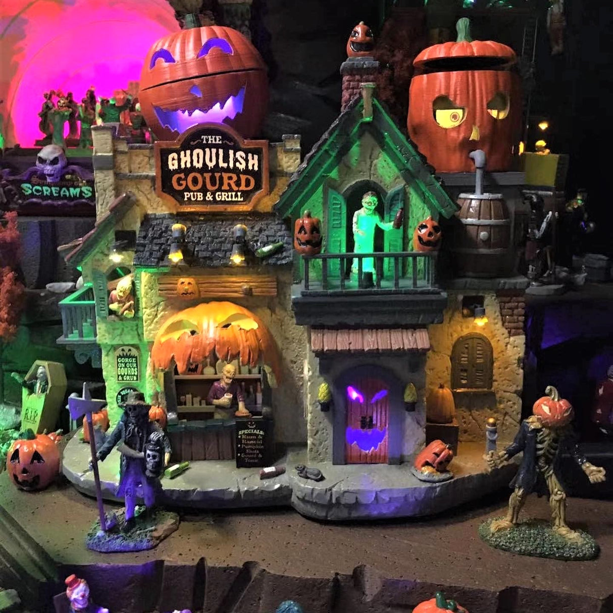 Polyresin lighted building for a Lemax Spooky Town display. The building looks like a rustic house. There are two animated jack-o-lanterns on the roof. There are lots of jack-o-lantern accents everywhere. The sign near the roof reads "THE GHOULISH GOURD PUB & GRILL." There are people on the balconies holding bottles. There's a person behind the counter.