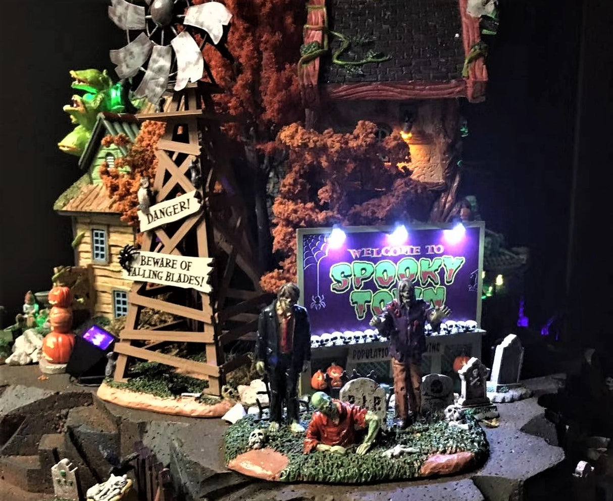 A Lemax Spooky Town display consisting of a spooky windmill, a cemetery with zombies, some tombstones, a pumpkin snowman, a spooky house, some orange trees and a lit up sign that reads "WELCOME TO SPOOKY TOWN!"