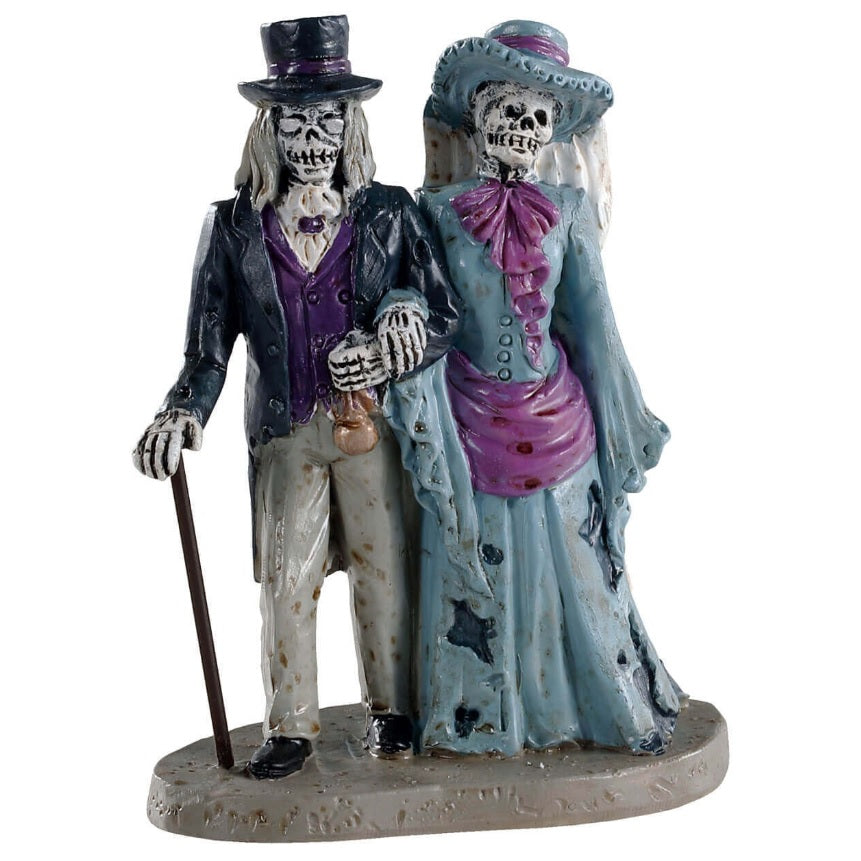 Polyresin figurine for a Lemax Spooky Town display. Designed to look like two skeletons dressed in raggedy, old Victorian-era style clothes. The man skeleton is wearing a suit and a top hat and walks with a cane. The woman skeleton is wearing a light blue and pink dress with a dirty, white veil.