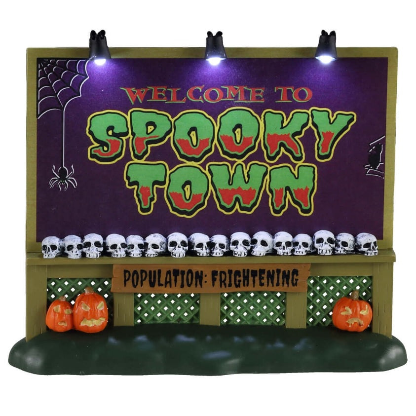 Plastic village accessory for a Lemax Spooky Town display. Designed to look like a town sign that reads "WELCOME TO SPOOKY TOWN." There are skulls lined up at the bottom of the sign. The sign also reads "POPULATION: FRIGHTENING." There are three jack-o-lanterns on the grass under the sign. The sign also has three lights on the top. The sign is purple, lime green, red, black, olive green and brown.