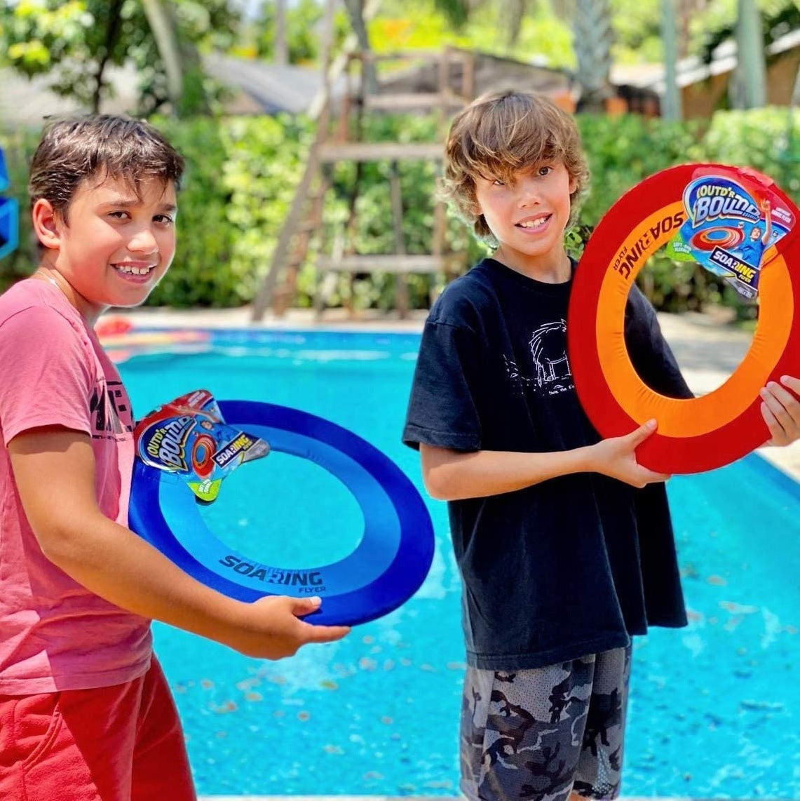 Lightweight frisbee made of colored spandex. There are two color options. One has a dark blue border, a light blue inner ring and the middle is missing so it flies further. The other one has a red border, an orange inner ring and the middle is missing so it flies further. This picture shows two boys holding them up next to a pool.