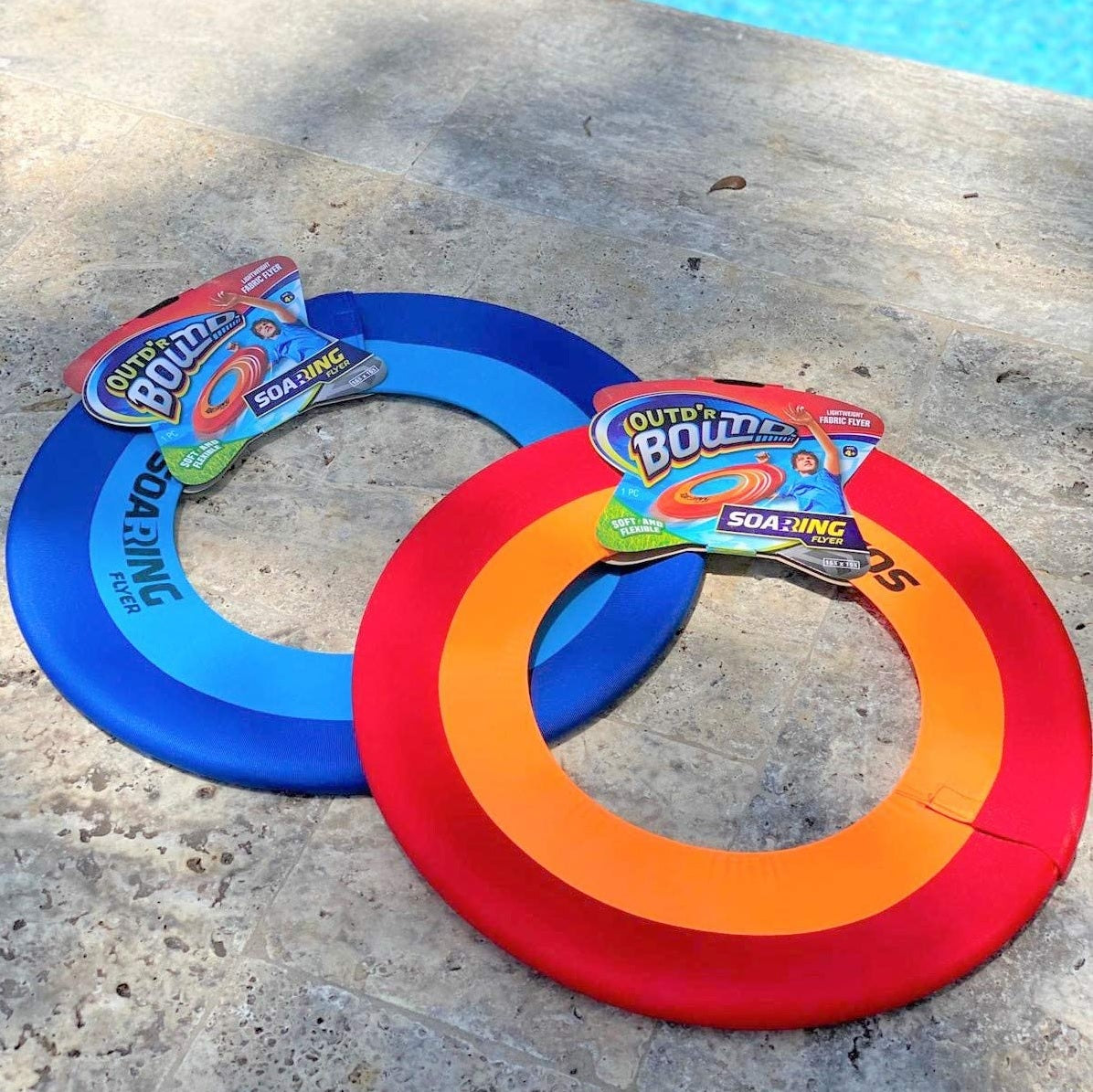 Lightweight frisbee made of colored spandex. There are two color options. One has a dark blue border, a light blue inner ring and the middle is missing so it flies further. The other one has a red border, an orange inner ring and the middle is missing so it flies further. This picture shows them on the concrete next to a pool.