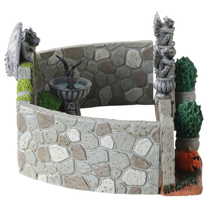 Polyresin village accent for a Lemax display. Designed to look like a stone fence with gargoyle accents. There's a birdbath with a crow in it and potted plants. There are two jack-o-lanterns on the ground near the entrance.