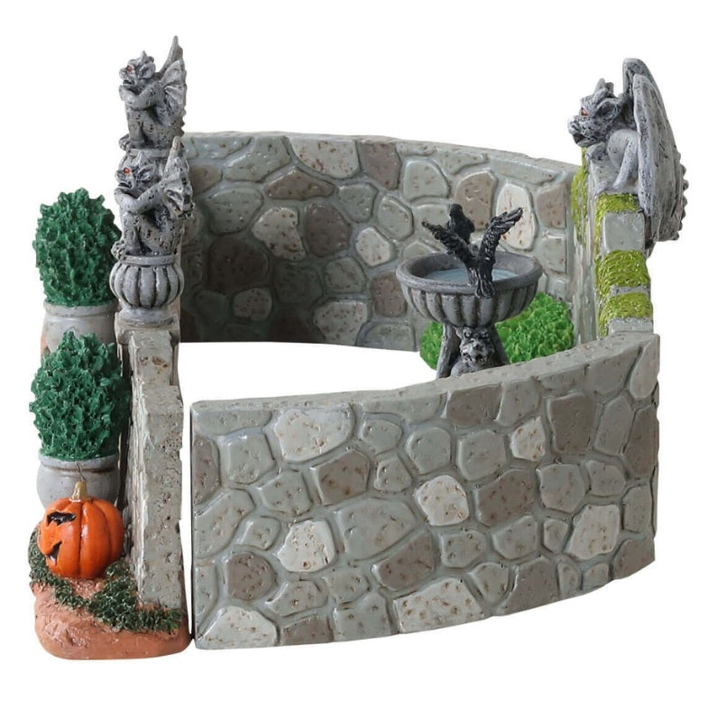 Polyresin village accent for a Lemax display. Designed to look like a stone fence with gargoyle accents. There's a birdbath with a crow in it and potted plants. There are two jack-o-lanterns on the ground near the entrance.