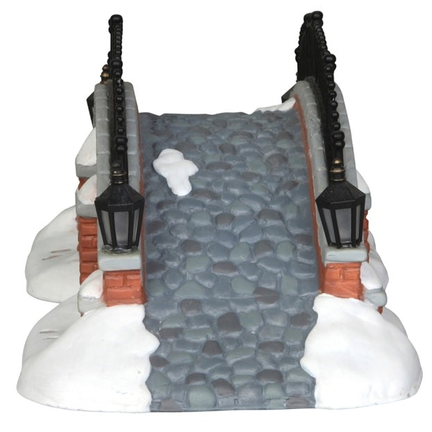 Porcelain lighted building for a Lemax display. Designed to look like a brick bridge with a metal fence. The path on the bridge is made of stone. There's snow on the ground and you can see footsteps in the snow.