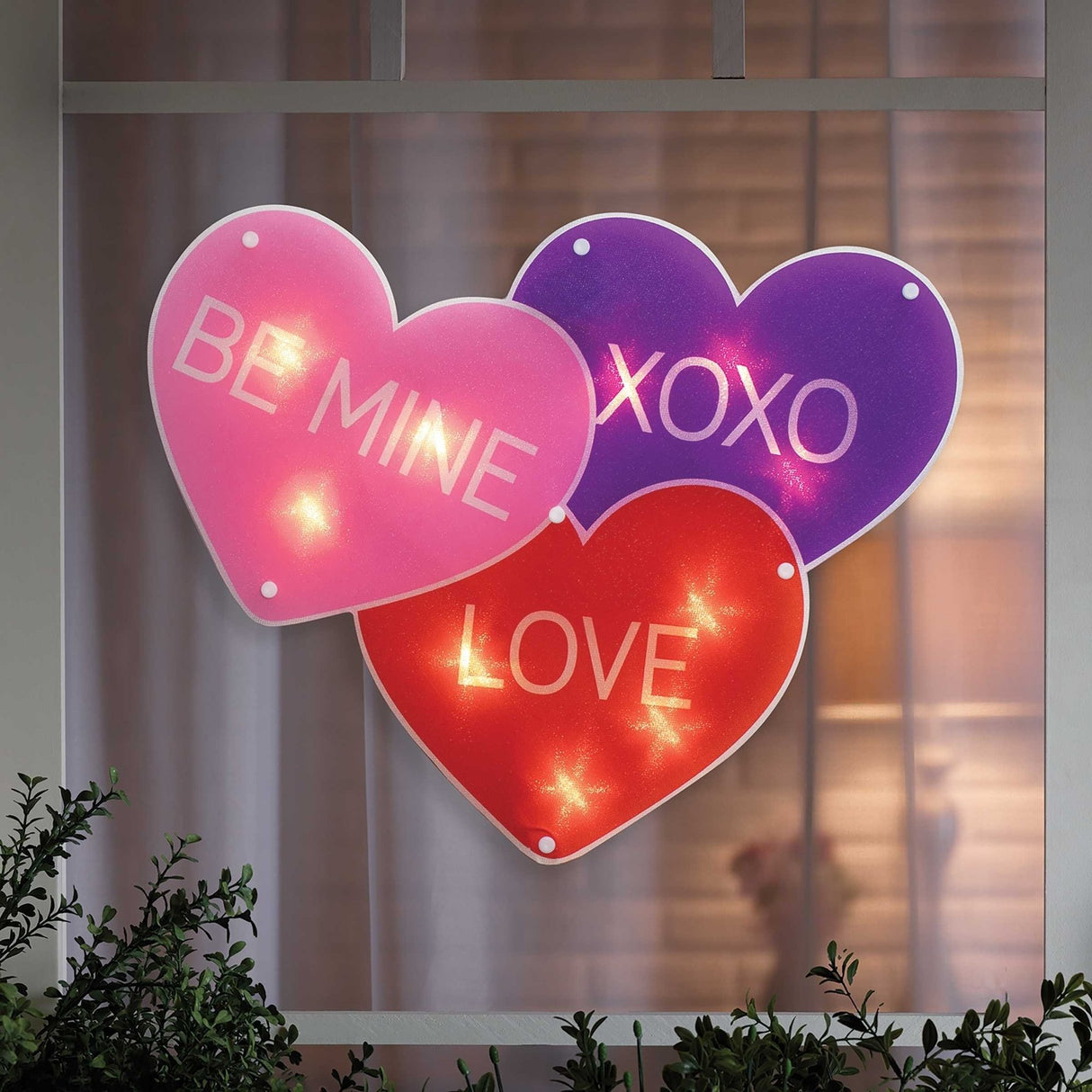 A light up decoration for Valentine's Day designed to look like three conversation hearts. One is purple and reads "XOXO." One is pink and reads "BE MINE." The other is red and reads "LOVE."