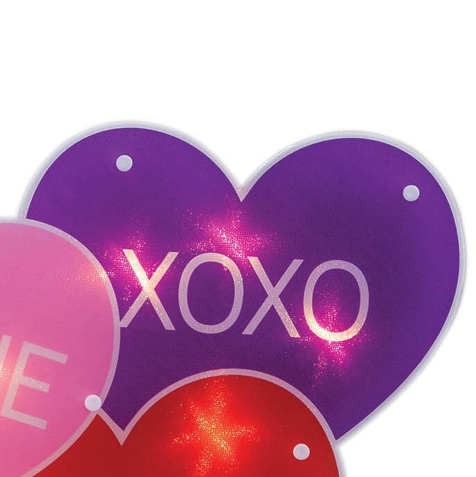 A light up decoration for Valentine's Day designed to look like three conversation hearts. One is purple and reads "XOXO." One is pink and reads "BE MINE." The other is red and reads "LOVE."
