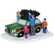 Polyresin figurine for a Lemax Village tabletop display. Designed to look like a family of four tying a Christmas tree to the top of their green and brown Vista Cruiser car. There's a small wreath with a red ribbon on the front bumper of the car. The ground is covered in snow. The young boy is holding a saw and the dad is wearing a Santa hat.