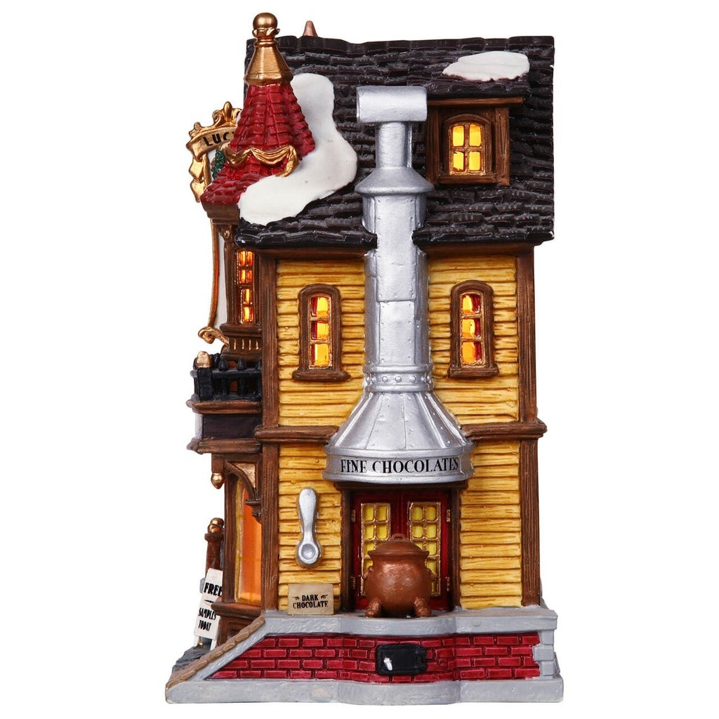 Porcelain lighted building for a Lemax display. Designed to look like an old Victorian-era house with wreaths and snow on it. The house is gold with brown frames. Inside the window you can see an array of chocolates.