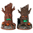Plastic village accessories for a Lemax display. Designed to look like two tree trunks with orange eyes and green, glowing mouths. They're sitting on grass and brick fences. One of the tree trunks is an orange-brown color and has a bunch of pumpkins near its base. The other is brown with a black cat and a pumpkin at its base.