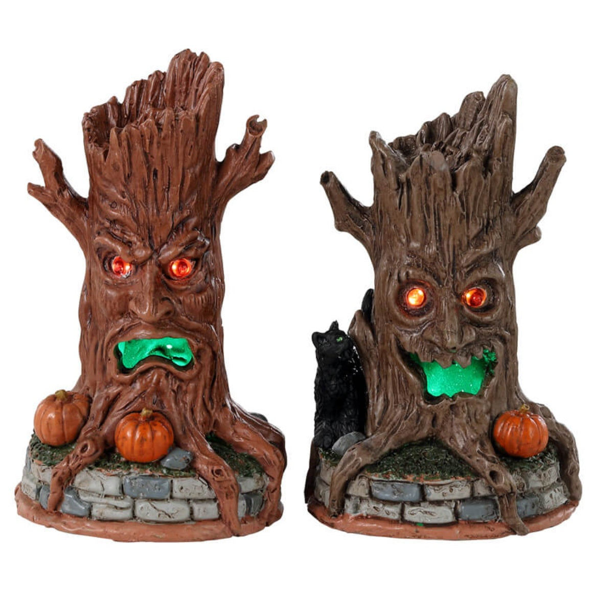 Plastic village accessories for a Lemax display. Designed to look like two tree trunks with orange eyes and green, glowing mouths. They're sitting on grass and brick fences. One of the tree trunks is an orange-brown color and has a bunch of pumpkins near its base. The other is brown with a black cat and a pumpkin at its base.