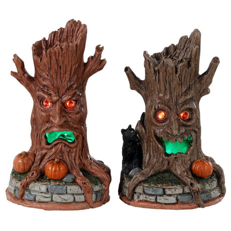 Plastic village accessories for a Lemax display. Designed to look like two tree trunks with orange eyes and green, glowing mouths. They're sitting on grass and brick fences. One of the tree trunks is an orange-brown color and has a bunch of pumpkins near its base. The other is brown with a black cat and a pumpkin at its base.