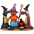 Resin figurine for a Lemax Spooky Town display. Designed to look like two witches building a snowman out of pumpkins. The pumpkin snowman is wearing a purple witch hat and a blue apron with a spider on it. The witch on the left is wearing a blue dress and the dress on the right side is wearing a violet dress. They're placing a broom in the hand of the pumpkin snowman. There's a pumpkin on the ground with a black cat inside.