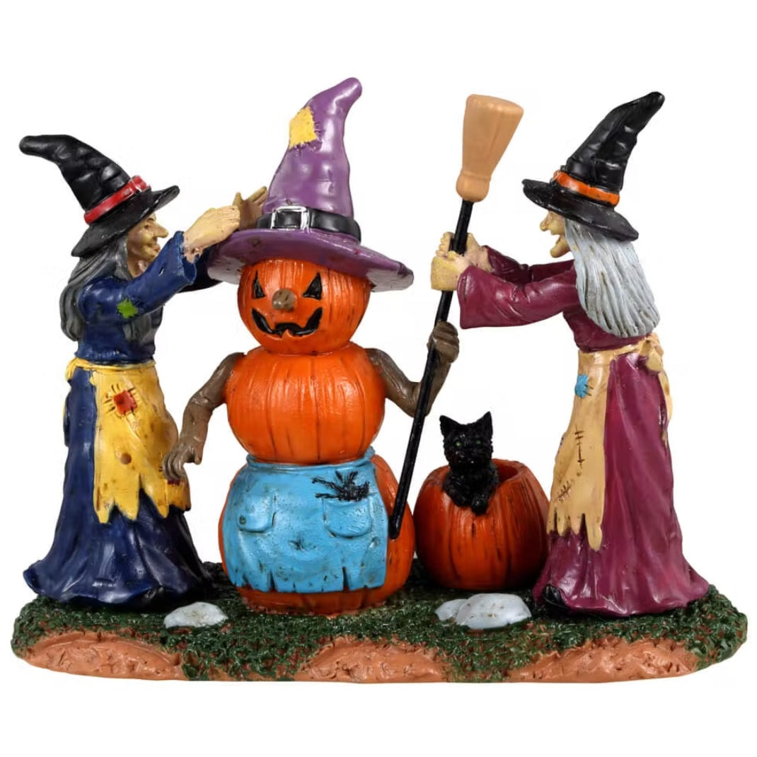Resin figurine for a Lemax Spooky Town display. Designed to look like two witches building a snowman out of pumpkins. The pumpkin snowman is wearing a purple witch hat and a blue apron with a spider on it. The witch on the left is wearing a blue dress and the dress on the right side is wearing a violet dress. They're placing a broom in the hand of the pumpkin snowman. There's a pumpkin on the ground with a black cat inside.