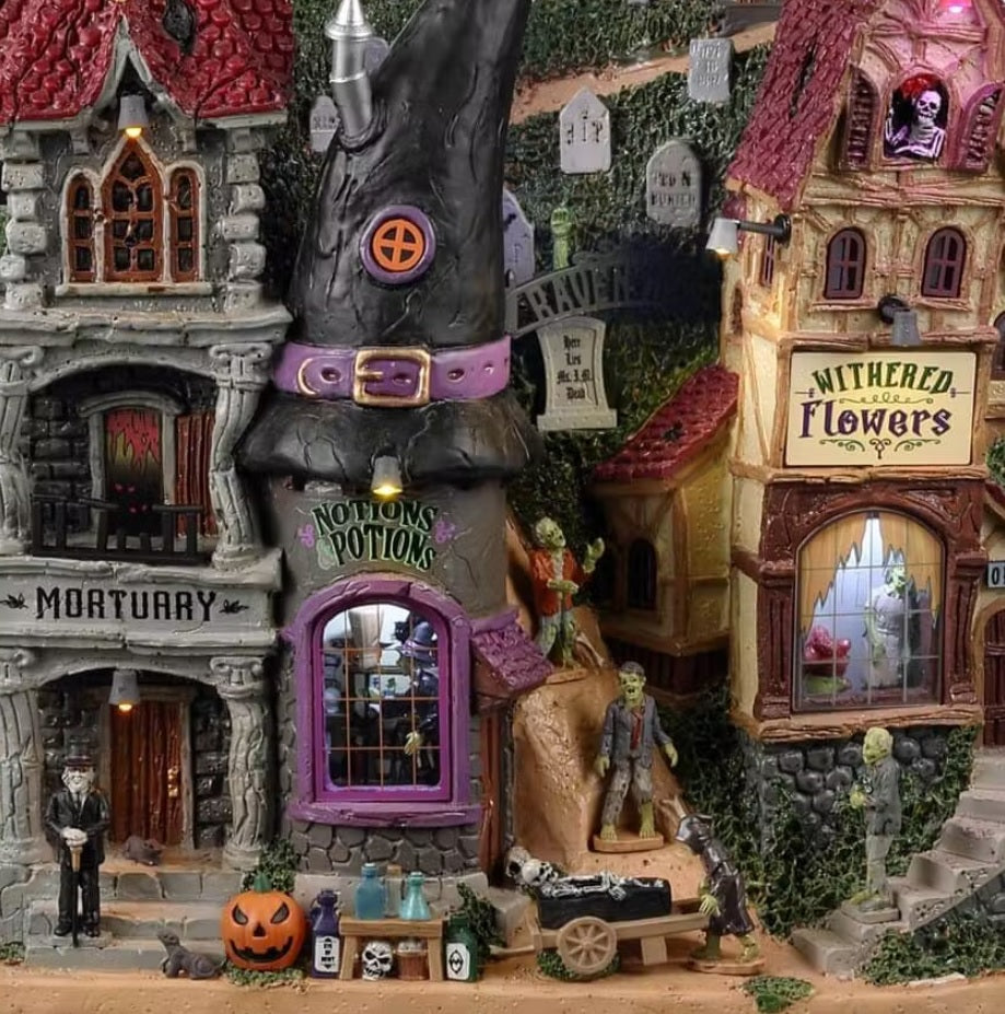 Resin building facade for a Spooky Town Lemax display. Designed to look like a bunch of store fronts. One section is a mortuary with a "help wanted" sign hanging from it. The building next to it reads "NOTION'S POTIONS" and has a witch hat for a roof. The building next to it is a flower shop. Behind them all is a cemetery and a spooky cathedral. There's a witch flying near the moon with bats following behind.