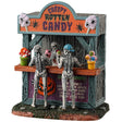 Resin village table accent for a Spooky Town Lemax display. Designed to look like a wooden candy stand. There's a skeleton serving candy to two young skeleton children. There are pots of candy on the side and pots of eyeballs on the other.