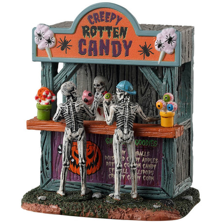 Resin village table accent for a Spooky Town Lemax display. Designed to look like a wooden candy stand. There's a skeleton serving candy to two young skeleton children. There are pots of candy on the side and pots of eyeballs on the other.
