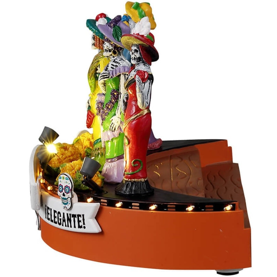 Resin accent for a Lemax Spooky Town display. Designed to look like five skeletons dressed in gaudy, brightly colored costumes. There are signs that read "Nuevo," "Dia de los Muertos Fashion Show" and "Elegante!" Designed to look like they're on stage at a fashion show. There are sugar skull accents and flowers on the stage.